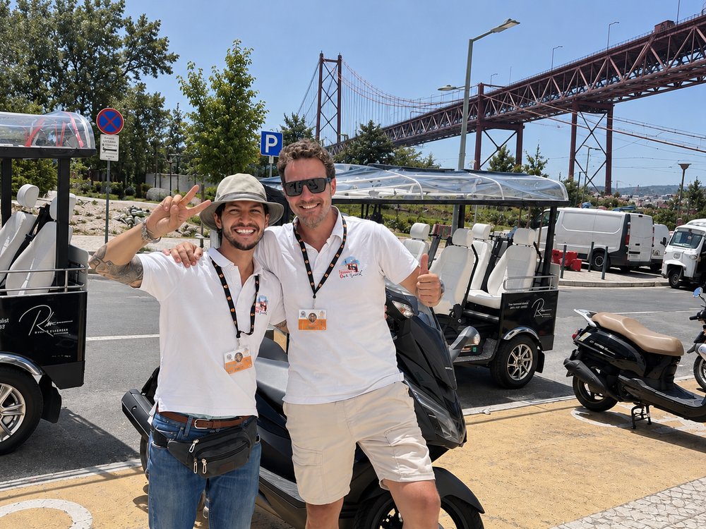 Two I Took a Tuk Tuk guides smiling, with the 25 de Abril bridge behind them